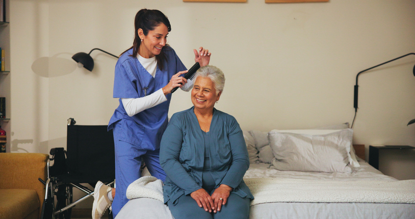 A cheerful caregiver in scrubs sits beside an older man on a couch, both engaged and smiling while interacting with a tablet. The scene highlights companionship, digital engagement, and supportive aged care in a comfortable, home-like setting.