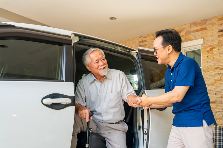 A cheerful caregiver in scrubs sits beside an older man on a couch, both engaged and smiling while interacting with a tablet. The scene highlights companionship, digital engagement, and supportive aged care in a comfortable, home-like setting.