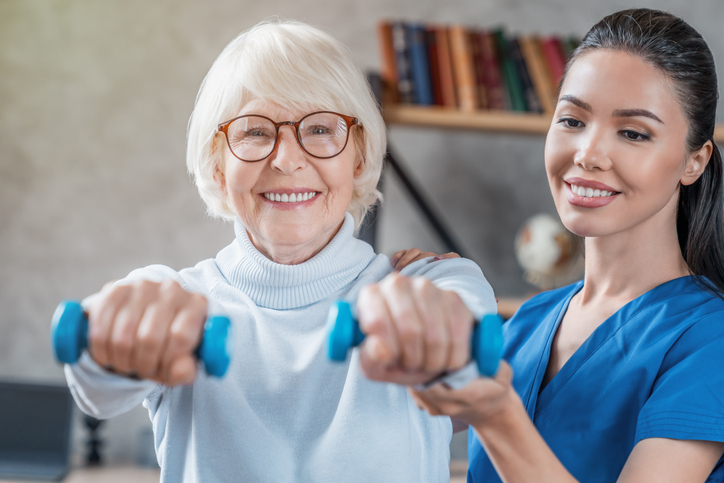 A cheerful caregiver in scrubs sits beside an older man on a couch, both engaged and smiling while interacting with a tablet. The scene highlights companionship, digital engagement, and supportive aged care in a comfortable, home-like setting.