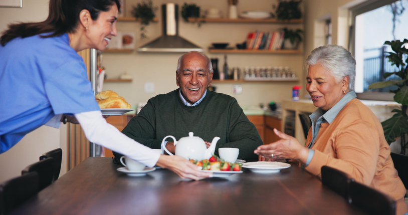 A cheerful caregiver in scrubs sits beside an older man on a couch, both engaged and smiling while interacting with a tablet. The scene highlights companionship, digital engagement, and supportive aged care in a comfortable, home-like setting.
