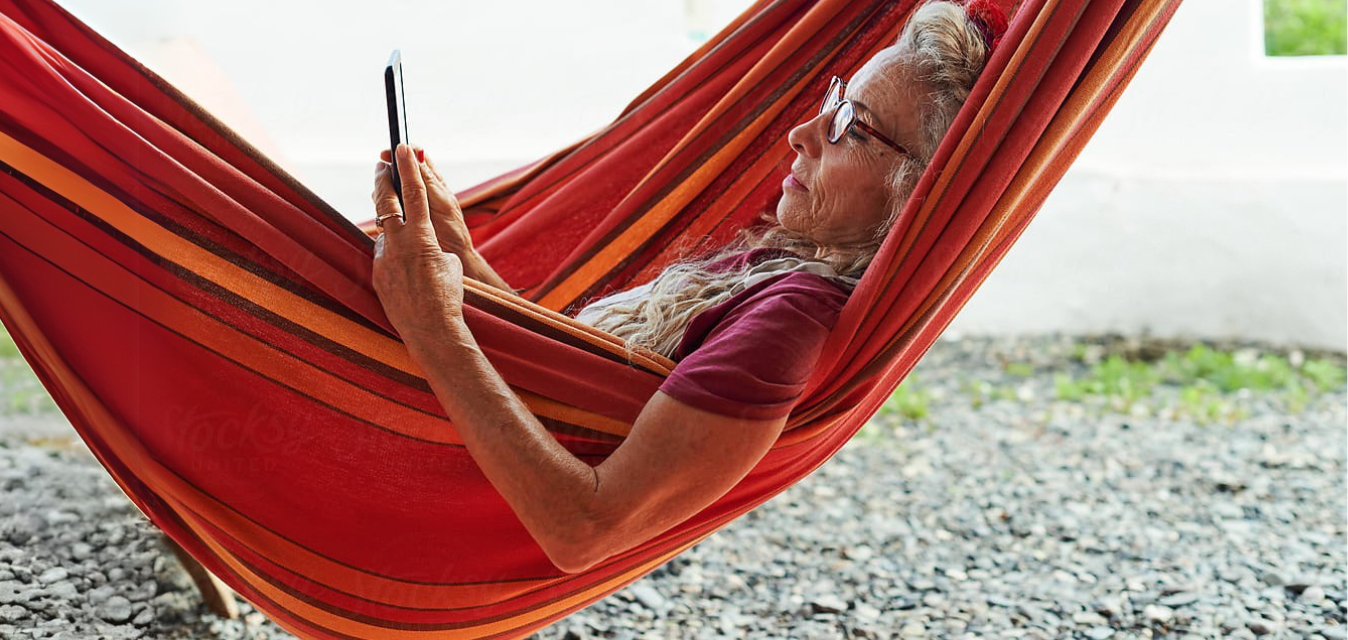 Elderly woman with grey hair and glasses relaxing in a red and orange hammock, reading a tablet.