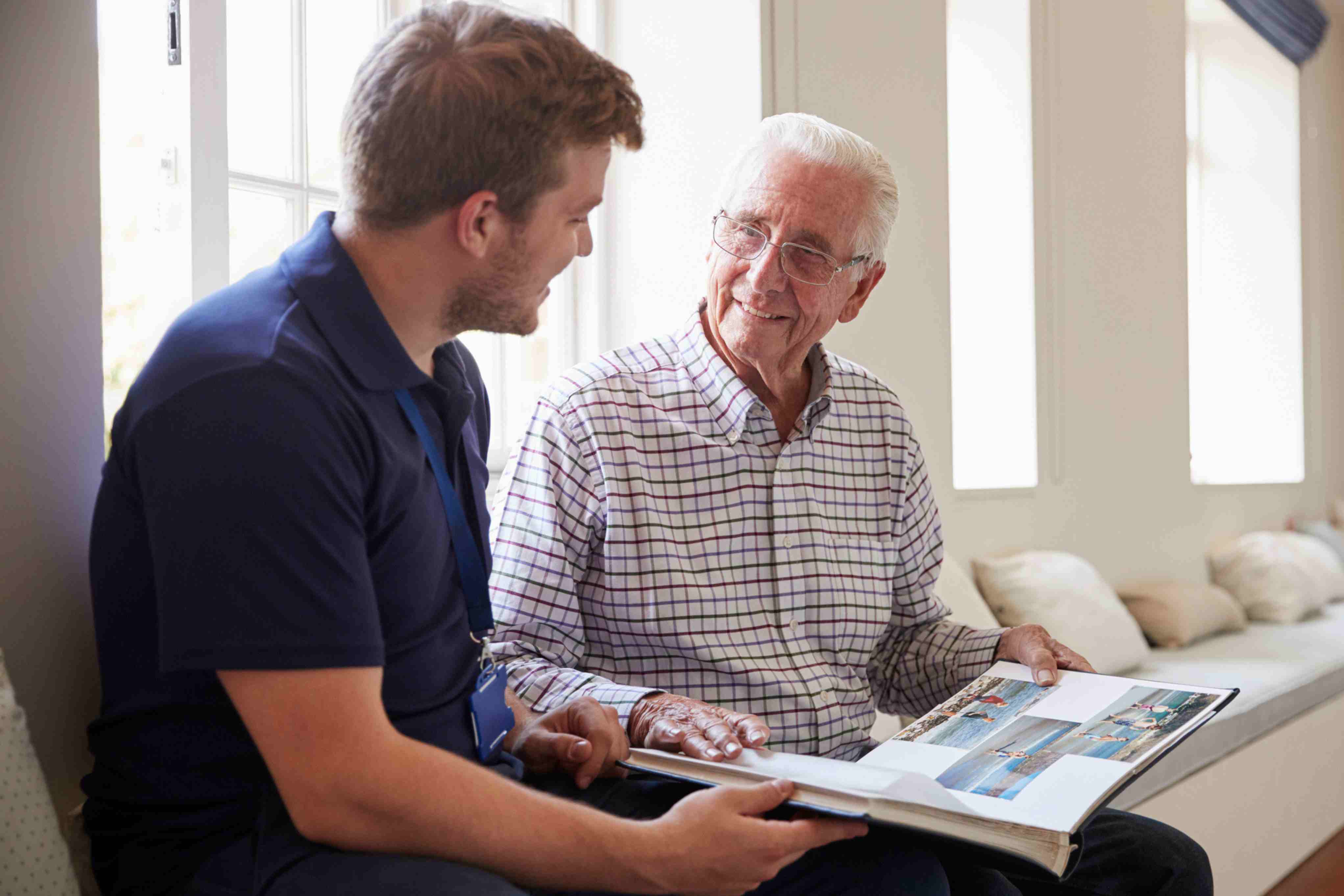 Caregiver sitting with an elderly man indoors, looking through a photo album together and sharing a warm conversation.