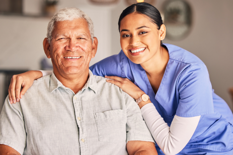 This image shows a smiling older man sitting beside a healthcare professional in scrubs, who has her arm gently around his shoulder. Both are looking at the camera warmly, conveying a sense of trust, care, and companionship.