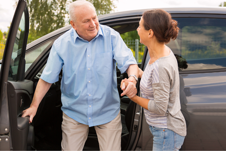 A caregiver assists an older man as he steps out of a car, suggesting transport or mobility support — reflecting independence and everyday assistance.