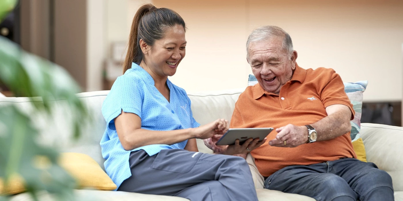 A smiling aged care worker sitting on a couch with an elderly man, both looking at a tablet together in a home-like setting.